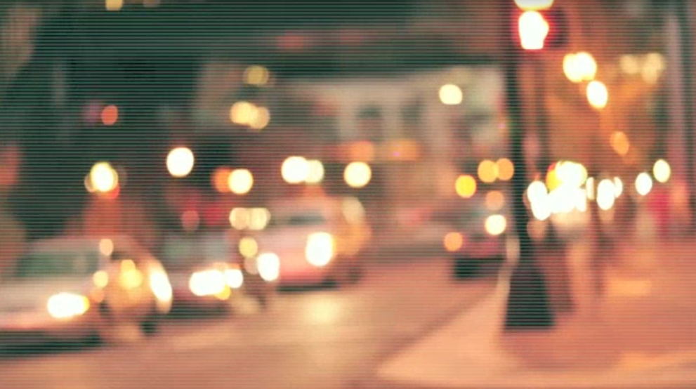 A dark urban street at night with soft yellow streetlights and warm bokeh in the distance.